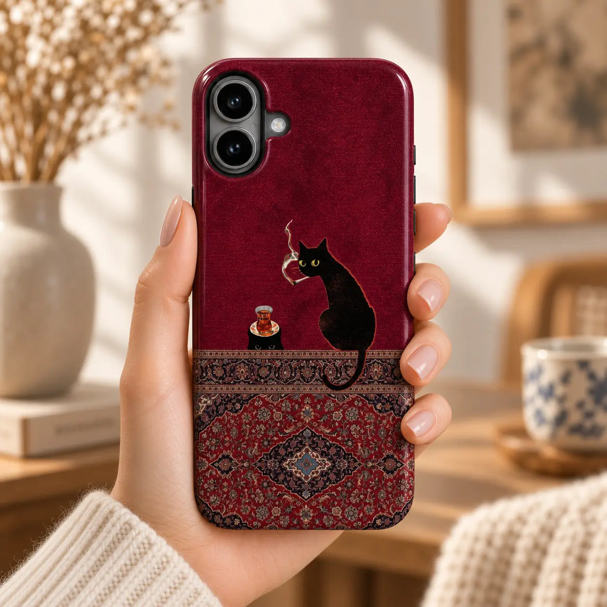Hand holding an iPhone with a burgundy case featuring black cats, a tea glass and an ornate Persian rug design