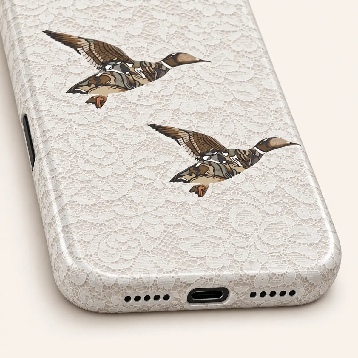 iPhone bottom close-up showing charging and speaker openings: white floral lace print; two camouflage ducks.