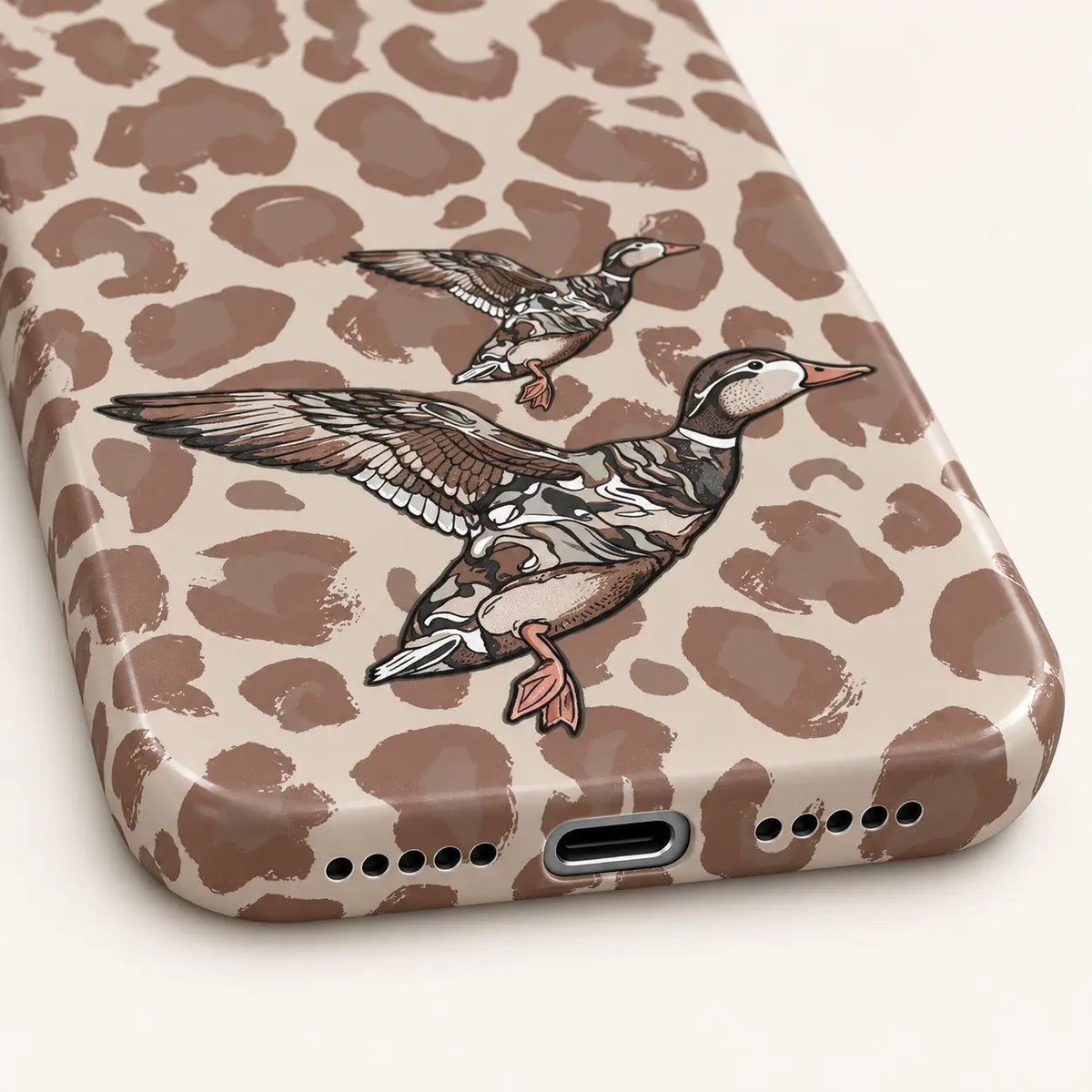 iPhone bottom close-up showing charging and speaker openings: beige and taupe leopard spots; two flying ducks, one large and one small.