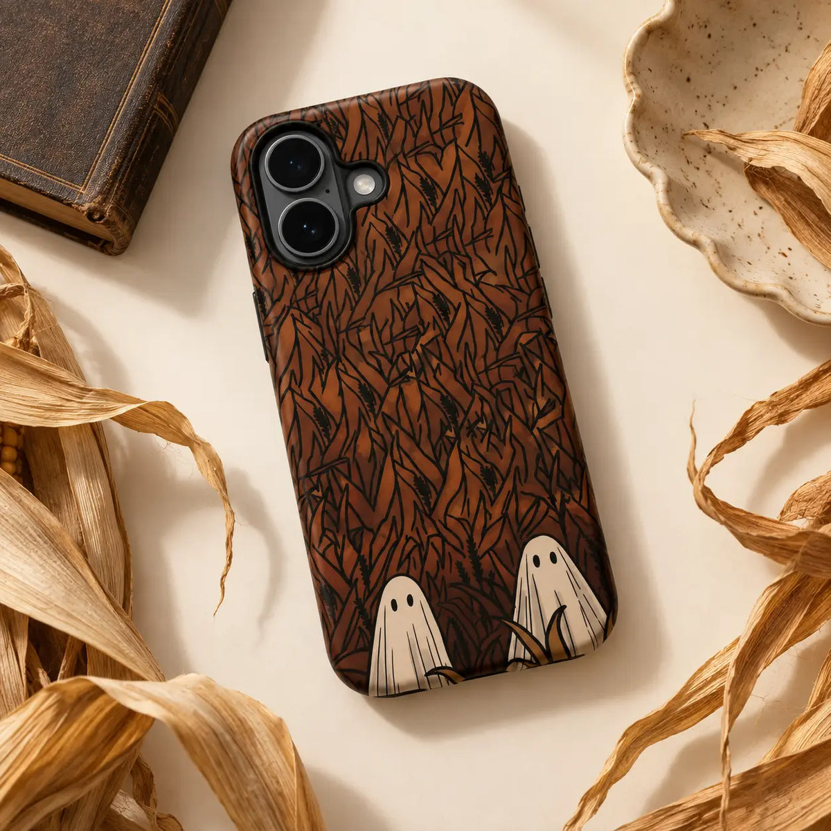 Hidden Ghosts cornfield phone case styled with dried corn leaves on a warm tabletop