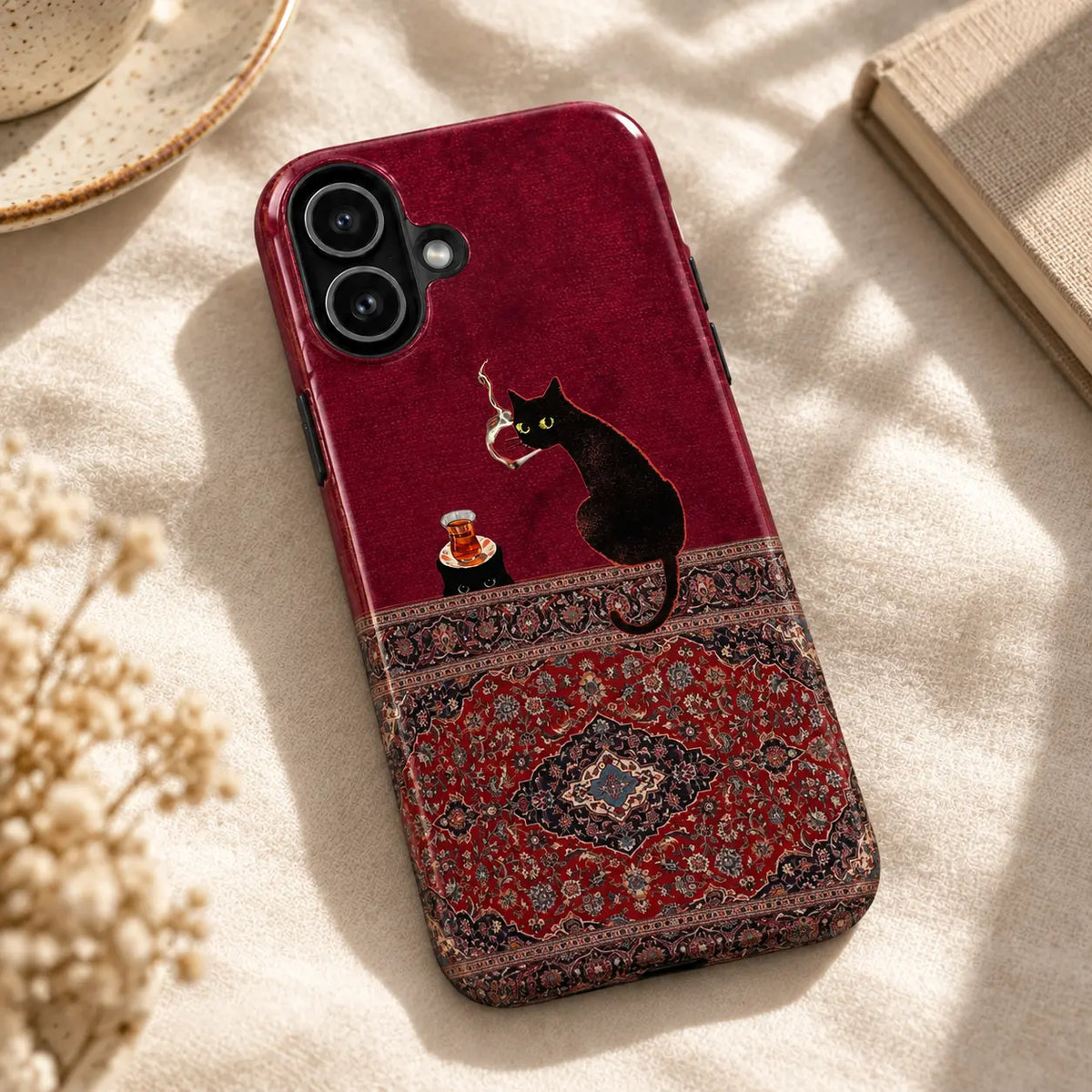 Burgundy black cat and Persian rug iPhone case resting on cream fabric beside a ceramic dish and a book