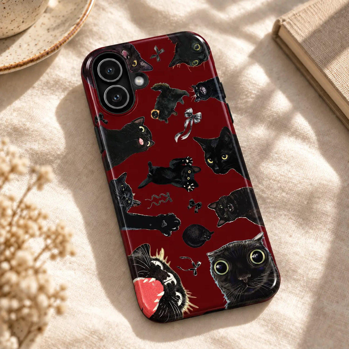 Burgundy black cat collage iPhone case resting on cream fabric beside a ceramic dish and a book