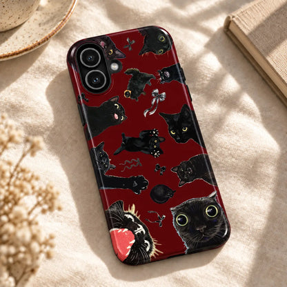 Burgundy black cat collage iPhone case resting on cream fabric beside a ceramic dish and a book