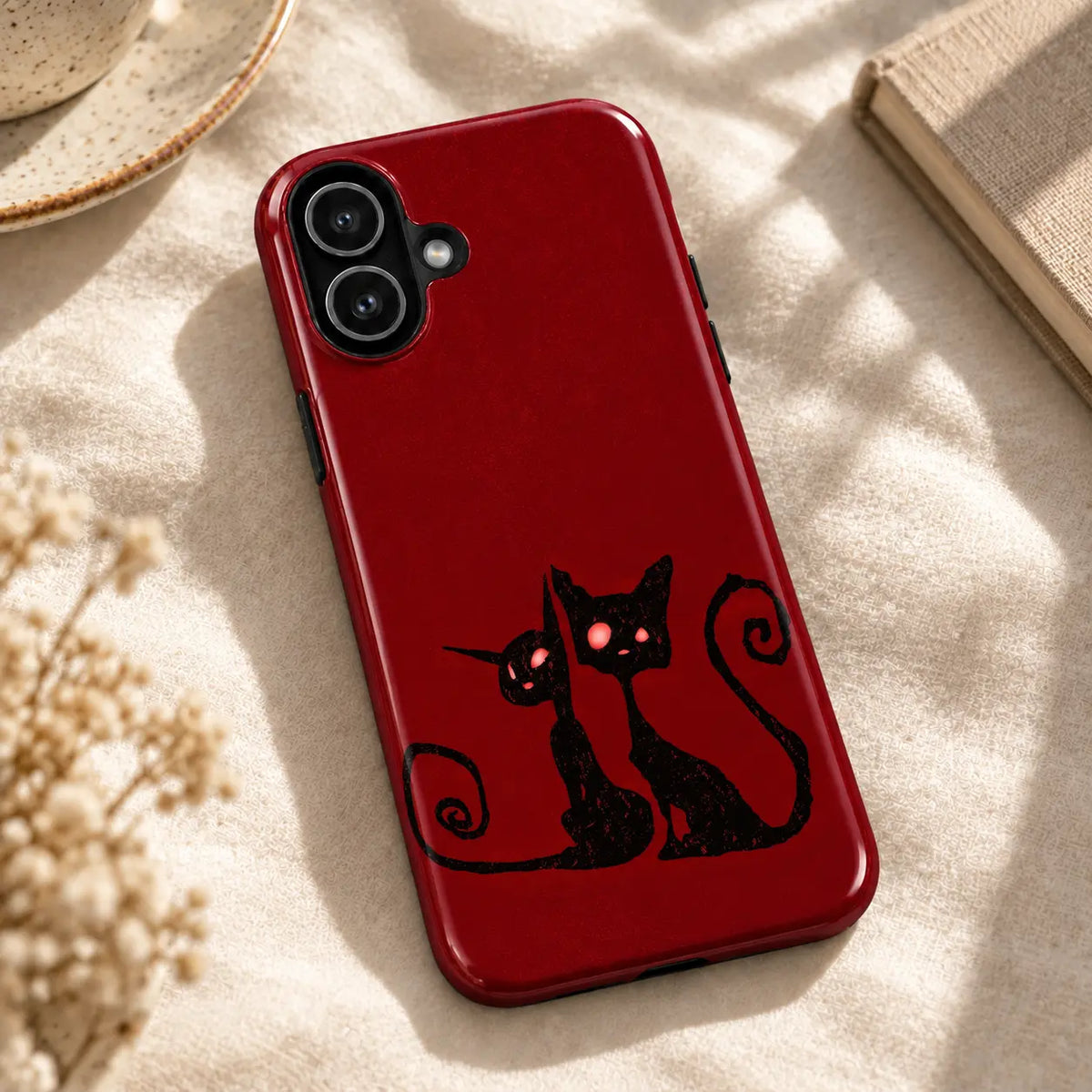 Crimson Cat Duo iPhone case resting on cream fabric beside a ceramic dish and a book