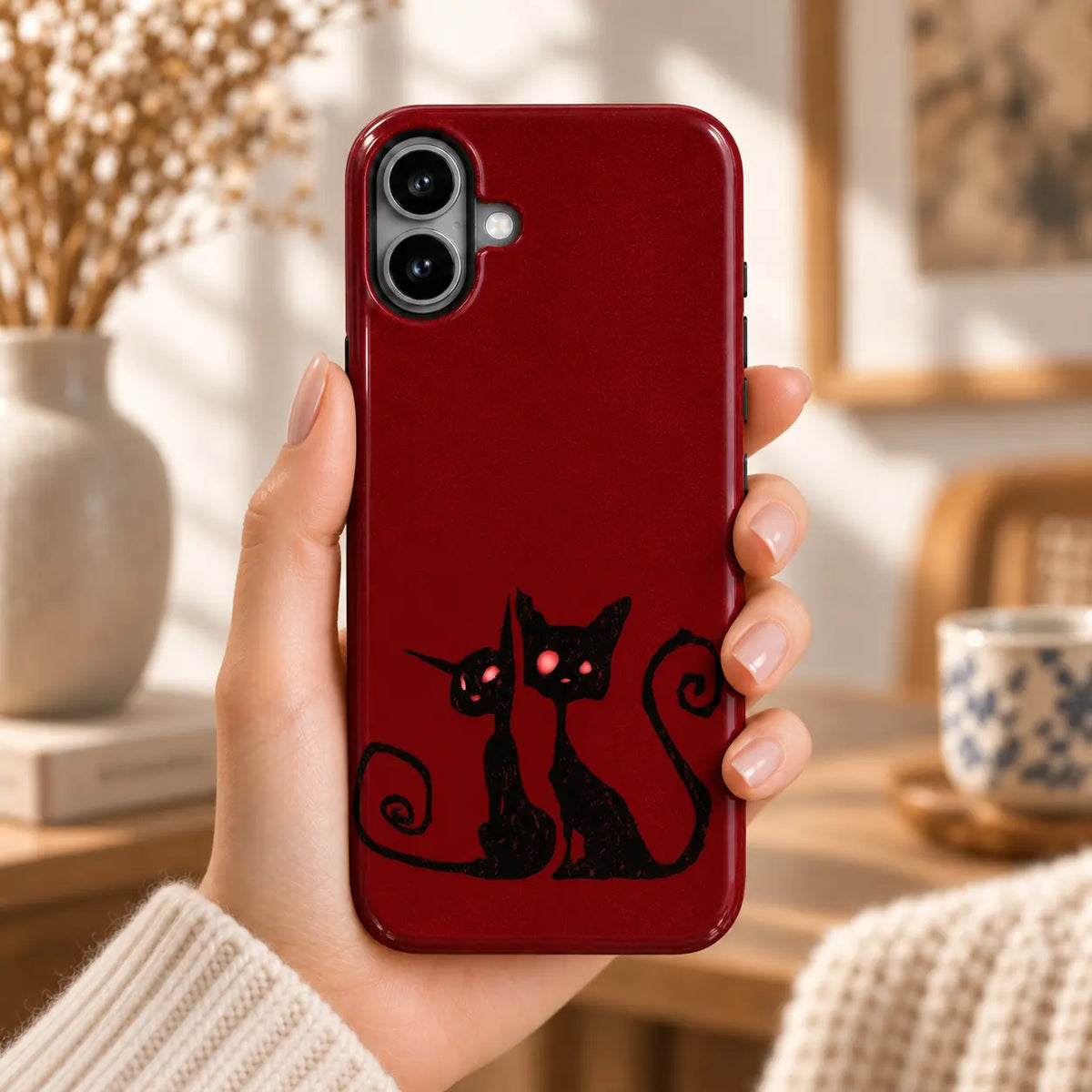Hand holding an iPhone with a crimson case featuring two black cats with curled tails and red eyes